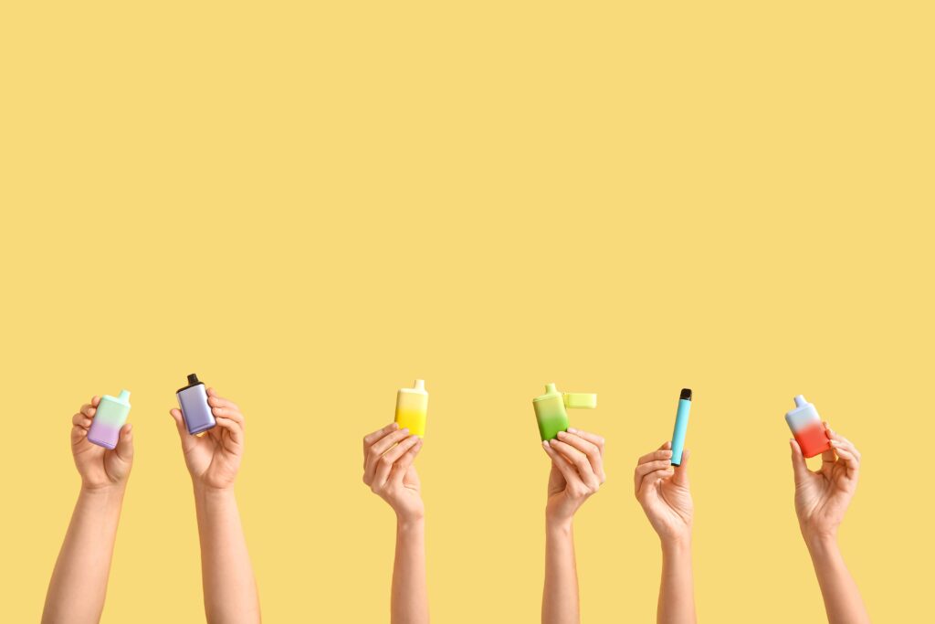 Mehrere Hände halten verschiedene bunte Einweg-E-Zigaretten vor einem gelben Hintergrund. Jede E-Zigarette hat eine unterschiedliche Farbe und Form.