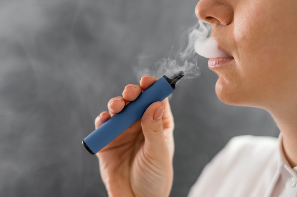 Eine Person hält eine blaue Einweg-E-Zigarette und dampft, während der Dampf aus ihrem Mund strömt. Nahaufnahme des Gesichts und der Hand vor grauem Hintergrund.